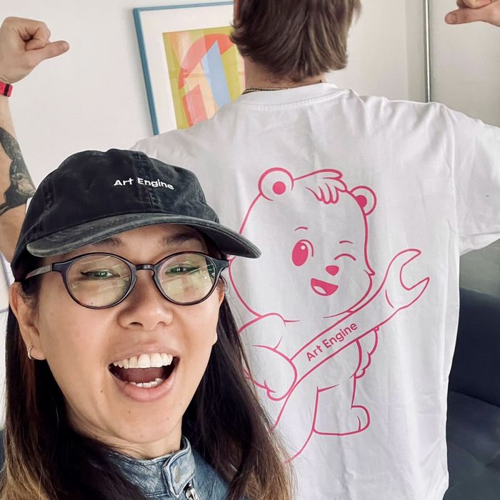 A smiling woman in an Art Engine cap, a friend behind her showing the winking bear on the back of a tee shirt