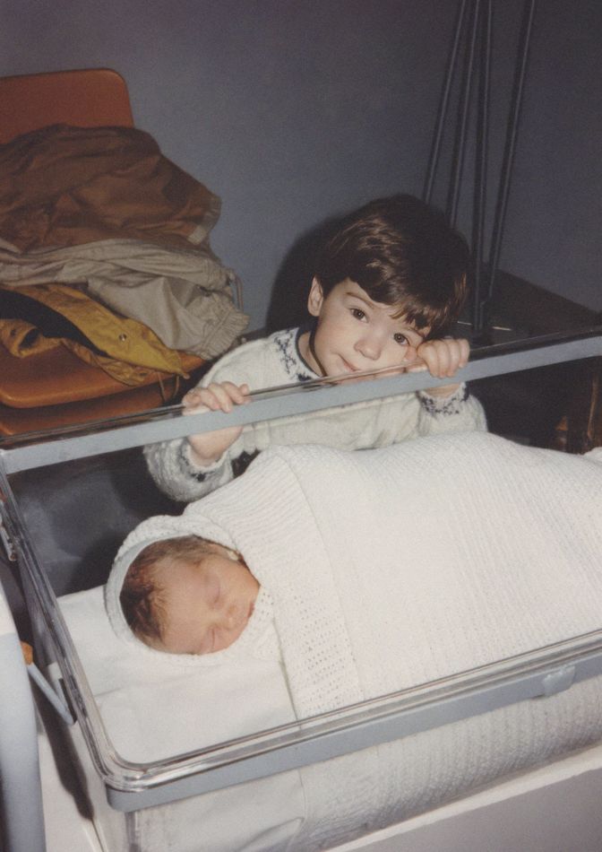 James as a toddler, peering into his newborn sibling&rsquo;s cot