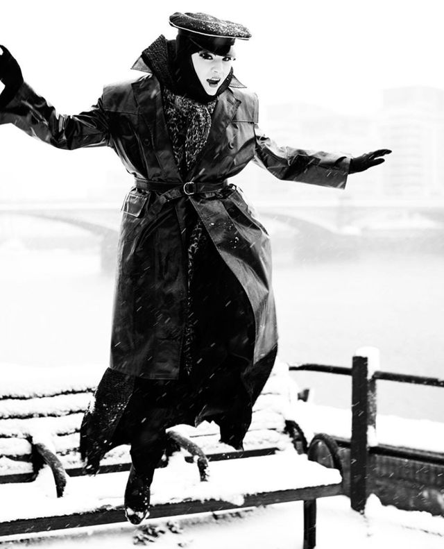 Daniel Lismore balancing on a snow-covered bench by the Thames in a long coat and cap, snow falling